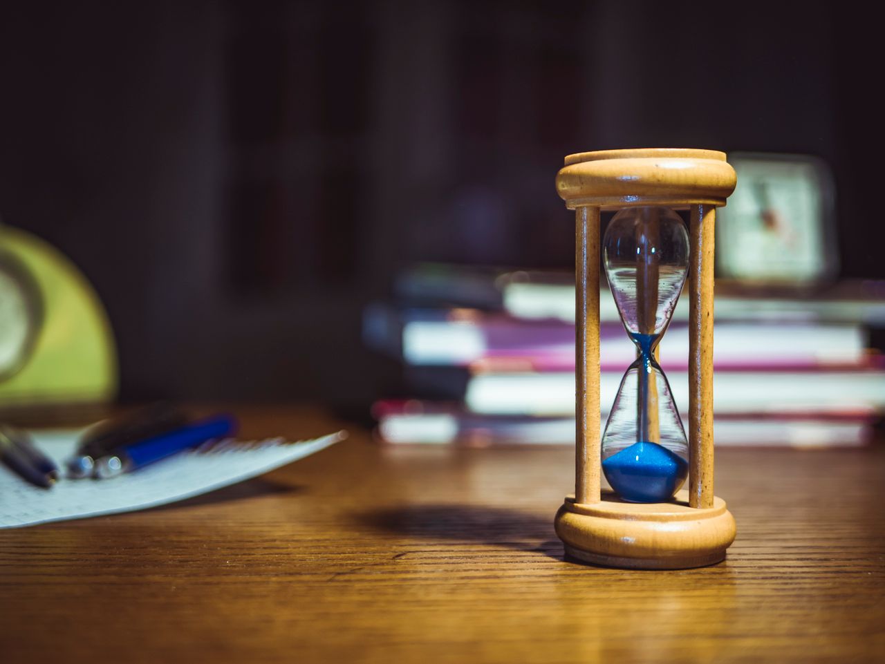 An hourglass in a brown frame on a wooden desk with books in background