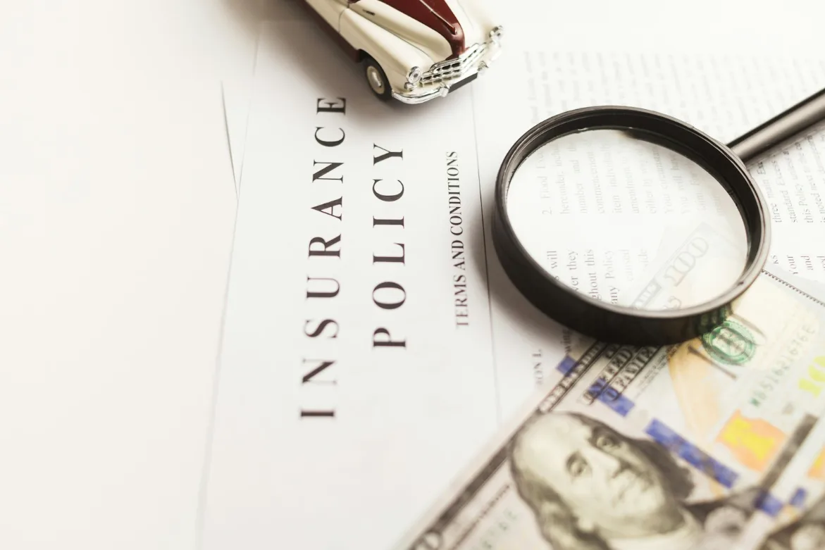 An insurance policy, terms and conditions document with a magnifying glass, 100 dollar bill, and toy car on top of it