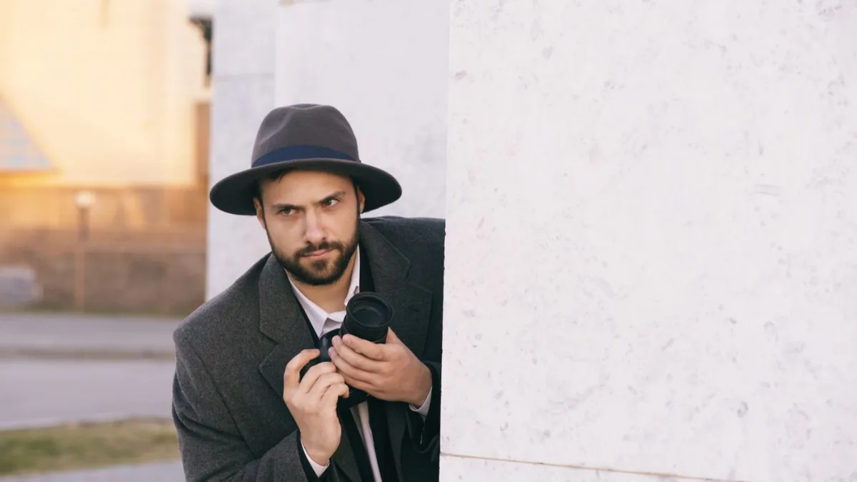 A man wearing a hat holding a camera peeking around corner A man wearing a hat holding a camera peeking around corner